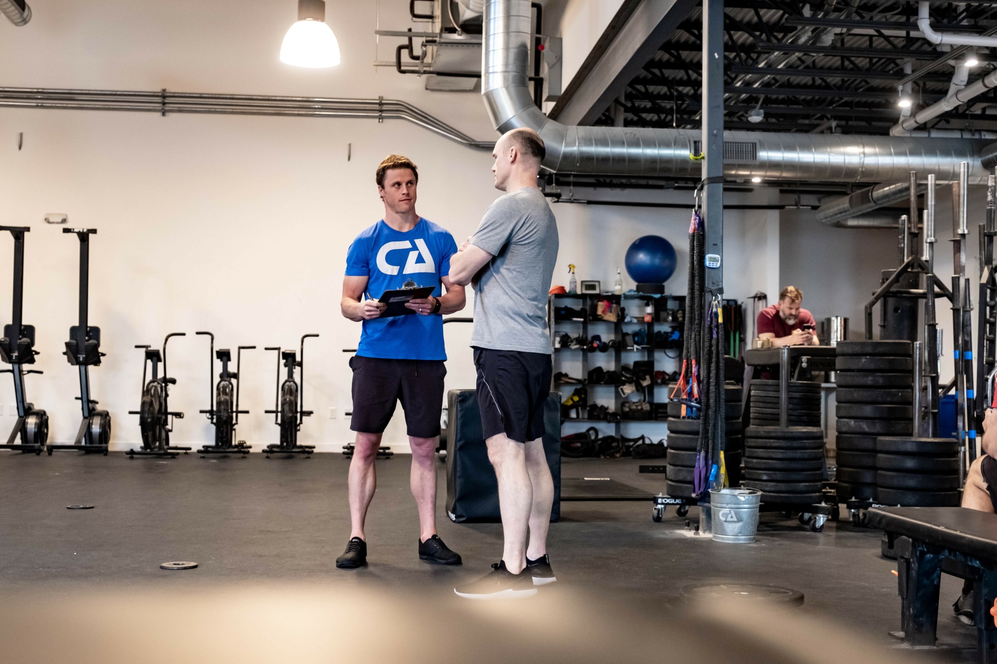 Two men stand and talk in a gym, one holding a phone, surrounded by workout equipment and weights.