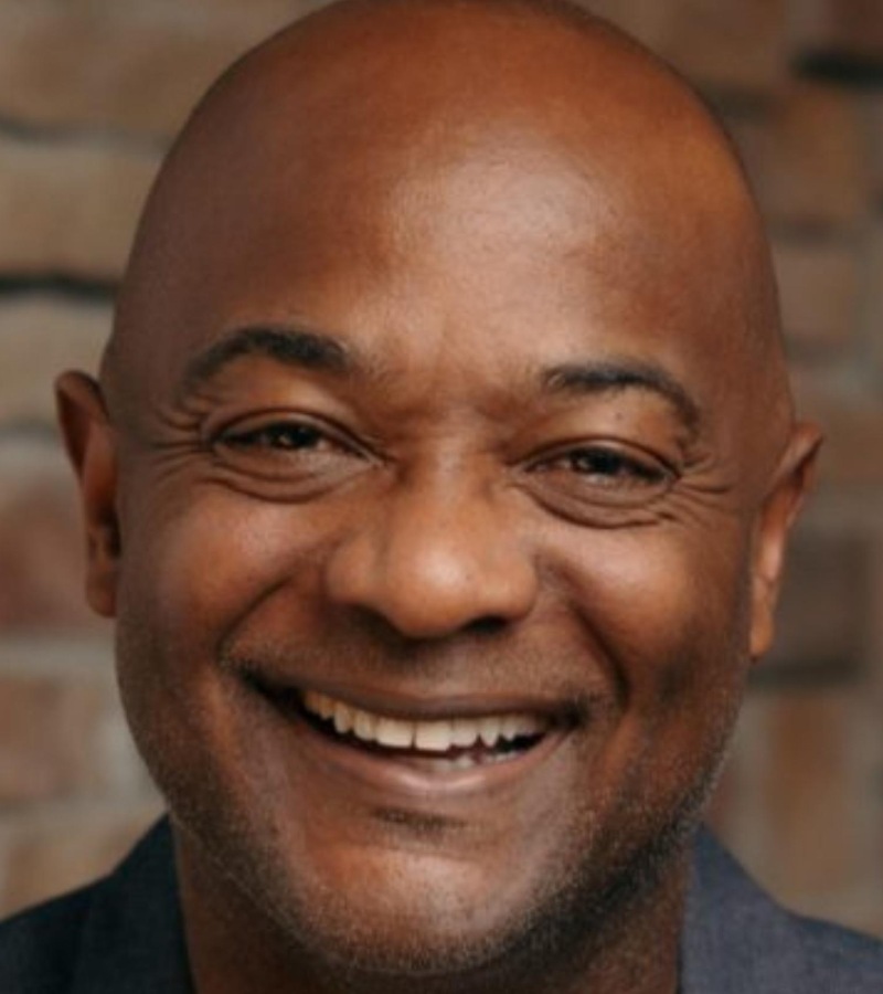 Close-up of smiling man with bald head and dark skin, background of brick wall.