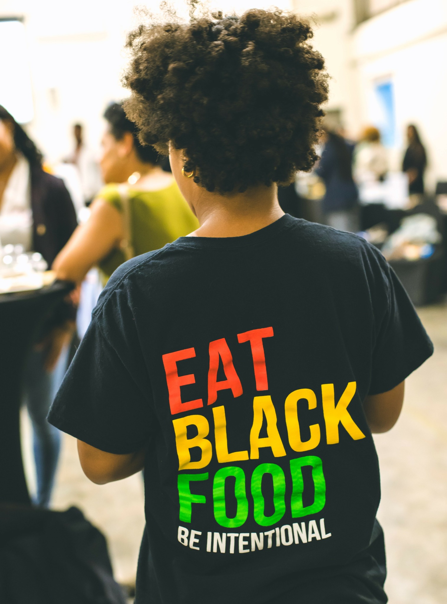 Person with curly hair wearing a black T-shirt with colorful text, in a busy outdoor setting.