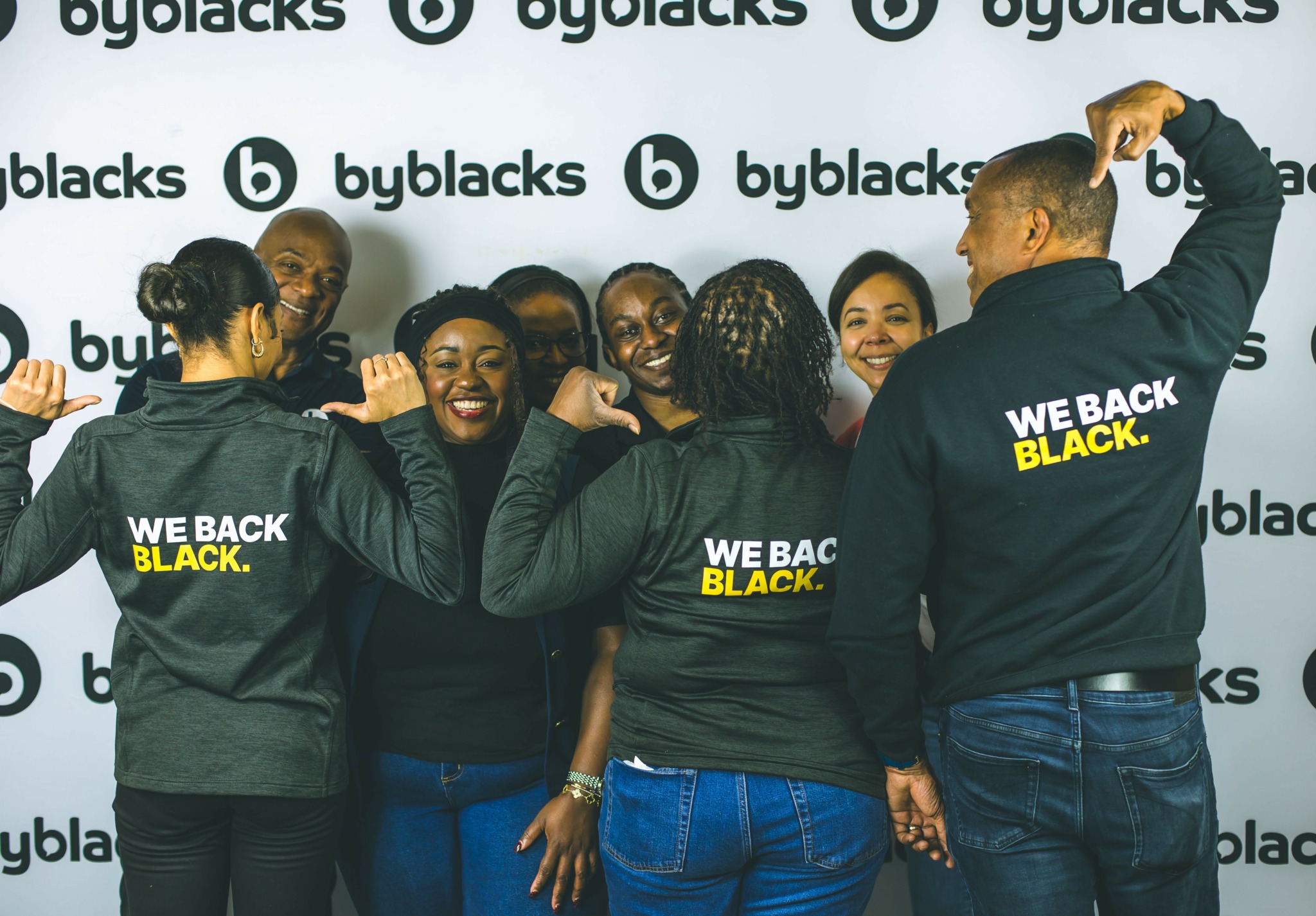Group of people wearing black jackets with yellow text, smiling and interacting in front of a backdrop with black and white logos.