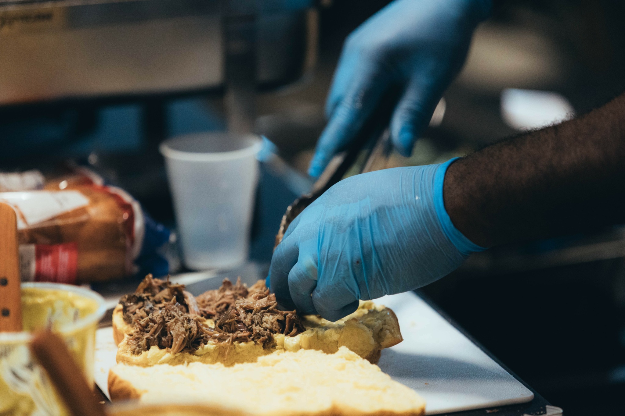 Person wearing blue gloves preparing food with hands on a cutting board, with various food items and a cup in background.