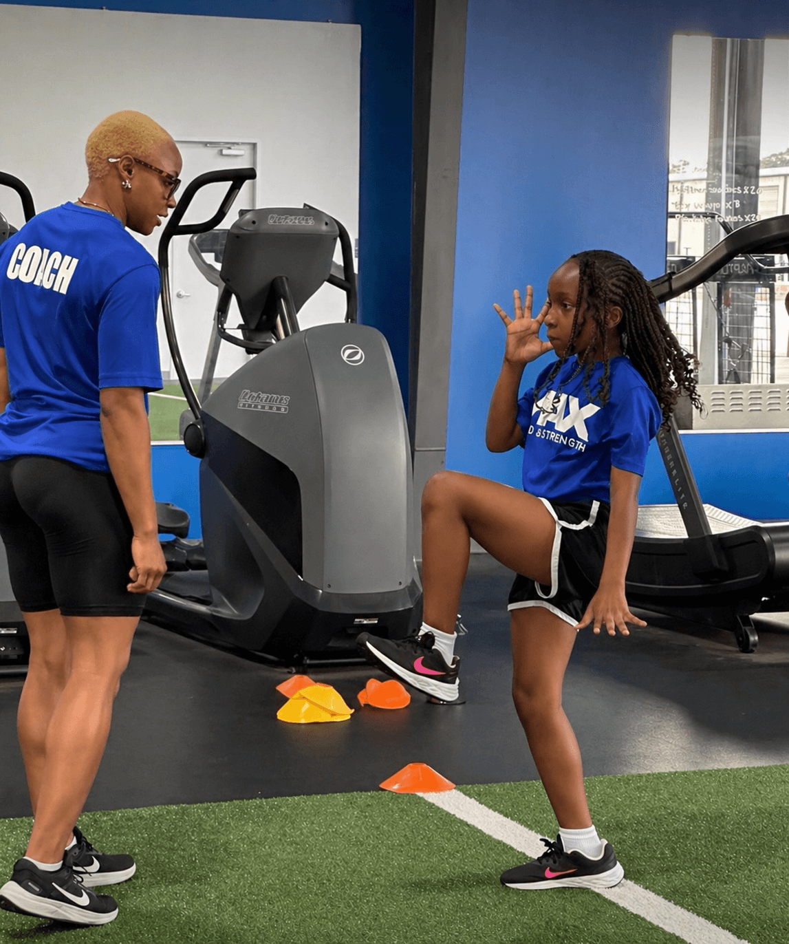 Girl stretching leg while trainer watches in gym with exercise equipment and blue walls.