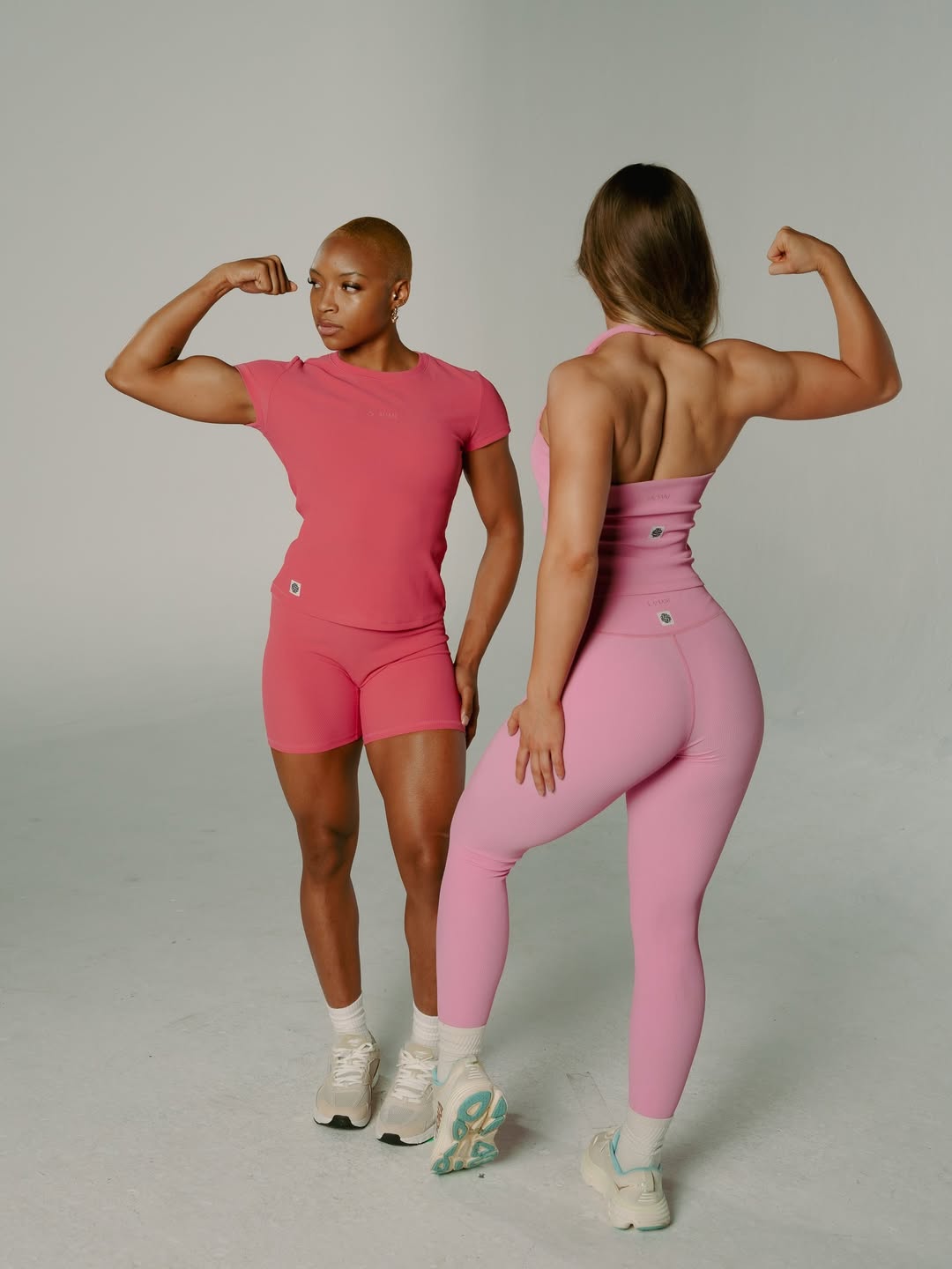 Two women in pink athletic outfits flexing muscles, facing away and to the side, against a plain background.