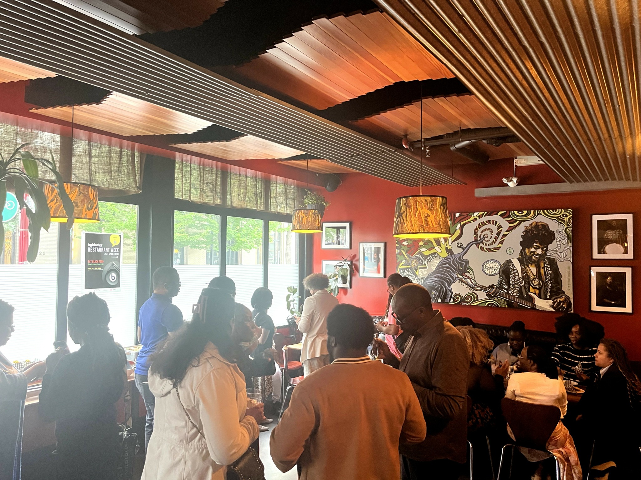 People gather inside a restaurant with large windows, wooden ceiling, and decorative wall art, some standing and talking.