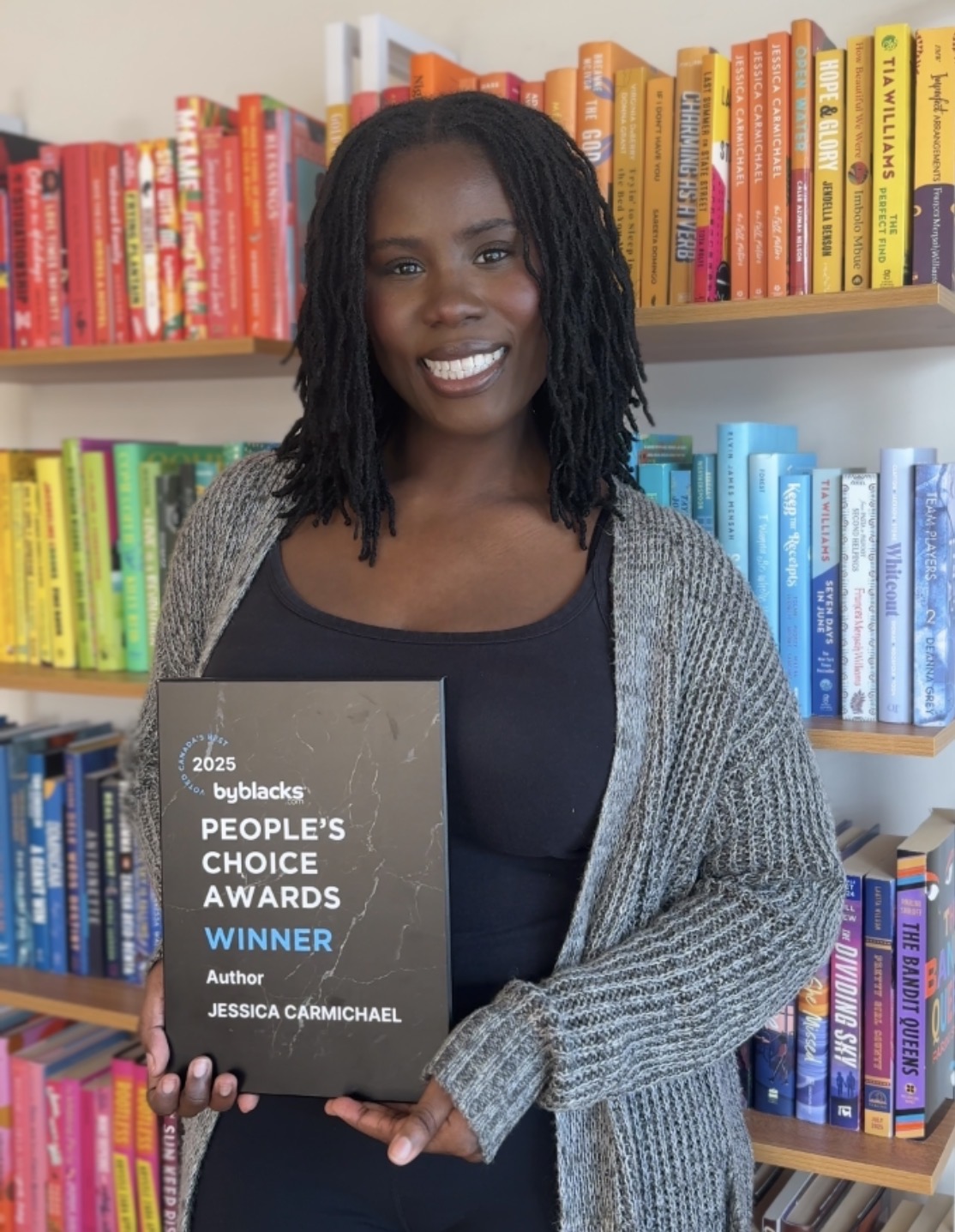 Woman holding award plaque in front of colorful bookshelf, smiling, wearing a gray cardigan and black top.