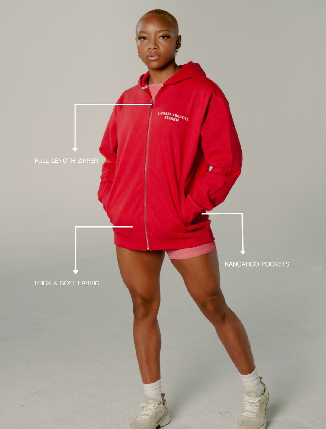 Woman in red hoodie and pink shorts, standing with hands in pockets, facing forward, neutral background.