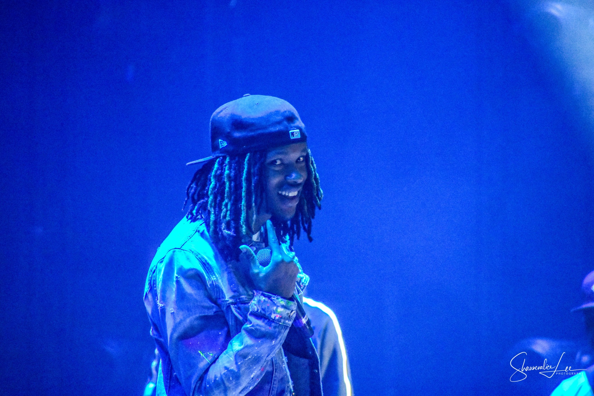 Person with dreadlocks wearing a cap and jacket, smiling, in a blue-lit environment.