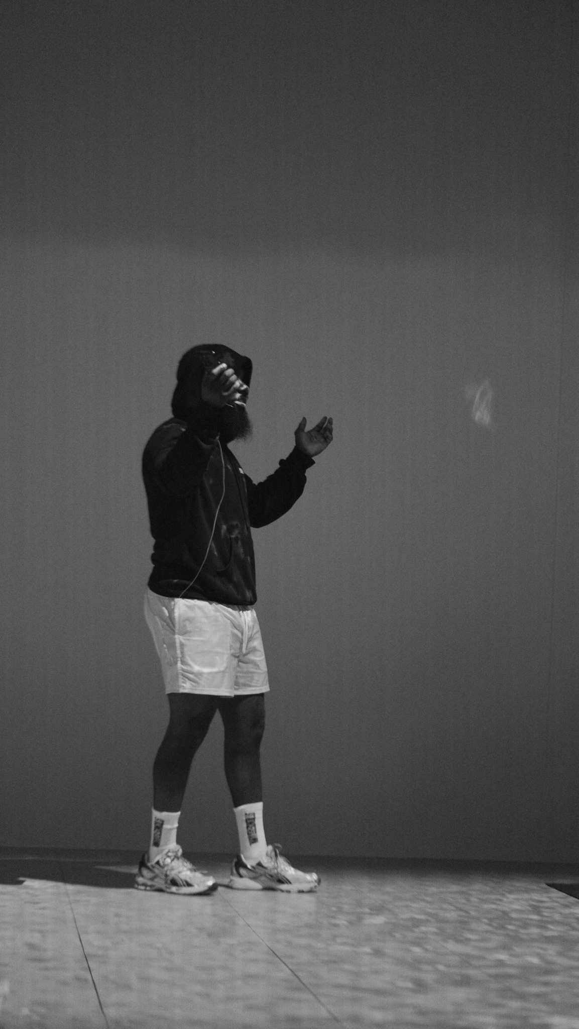 Person standing on stage, wearing shorts, sneakers, and a jacket, holding a microphone, against a plain background.
