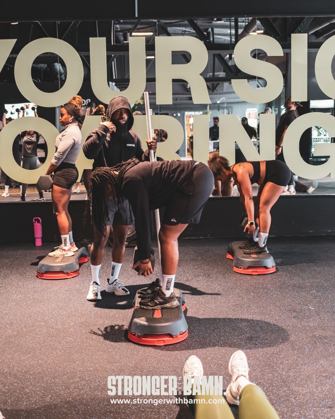People exercising with step platforms in a gym, large sign in background, some bending over, others standing.