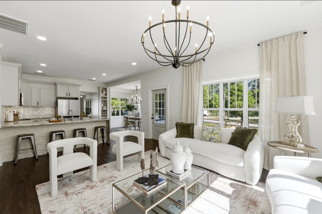 Bright living room with white sofa, chairs, glass coffee table, chandelier, and large windows with curtains.