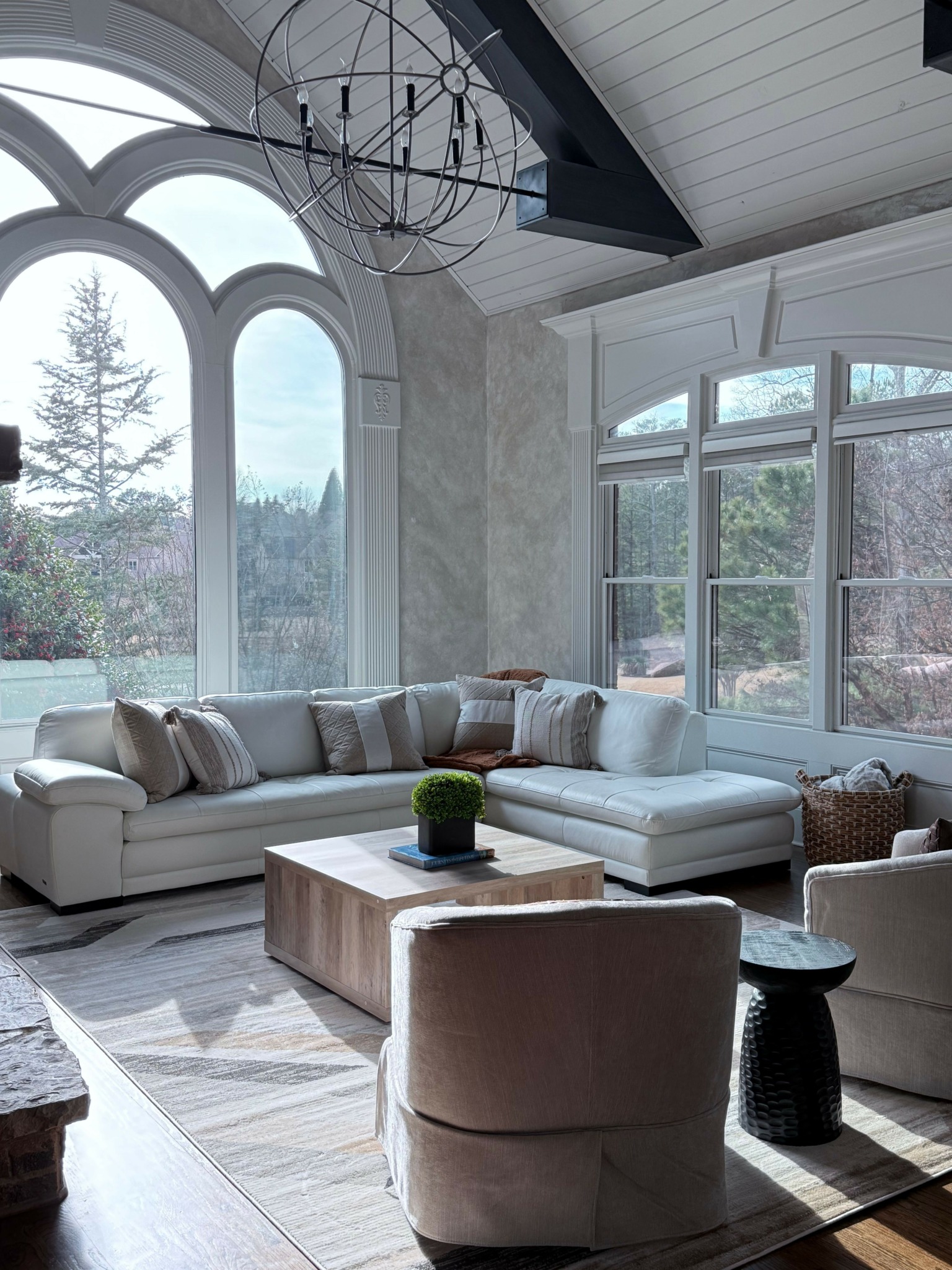 Living room with large window, white sofa, coffee table, and armchair, decorated with plants and a chandelier.