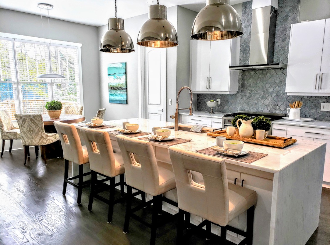 Modern kitchen with white cabinets, marble backsplash, island with seating, pendant lights, and a dining area with chairs and table.