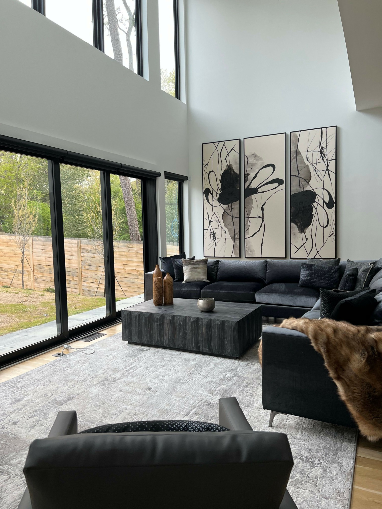 Living room with black sofa, artwork, and large windows showing greenery outside.