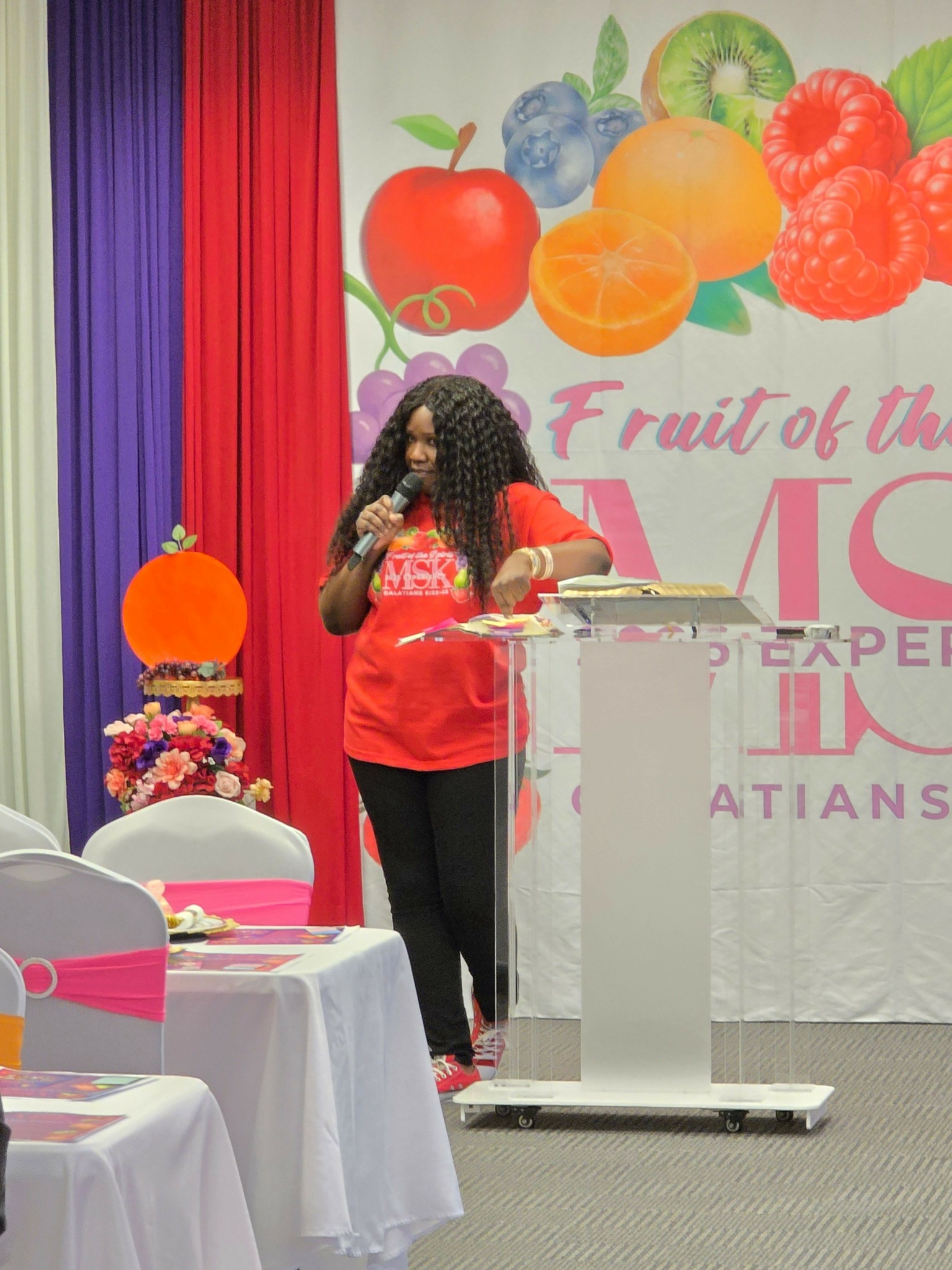 Woman standing at podium with colorful backdrop, table with cake, and chairs in foreground.