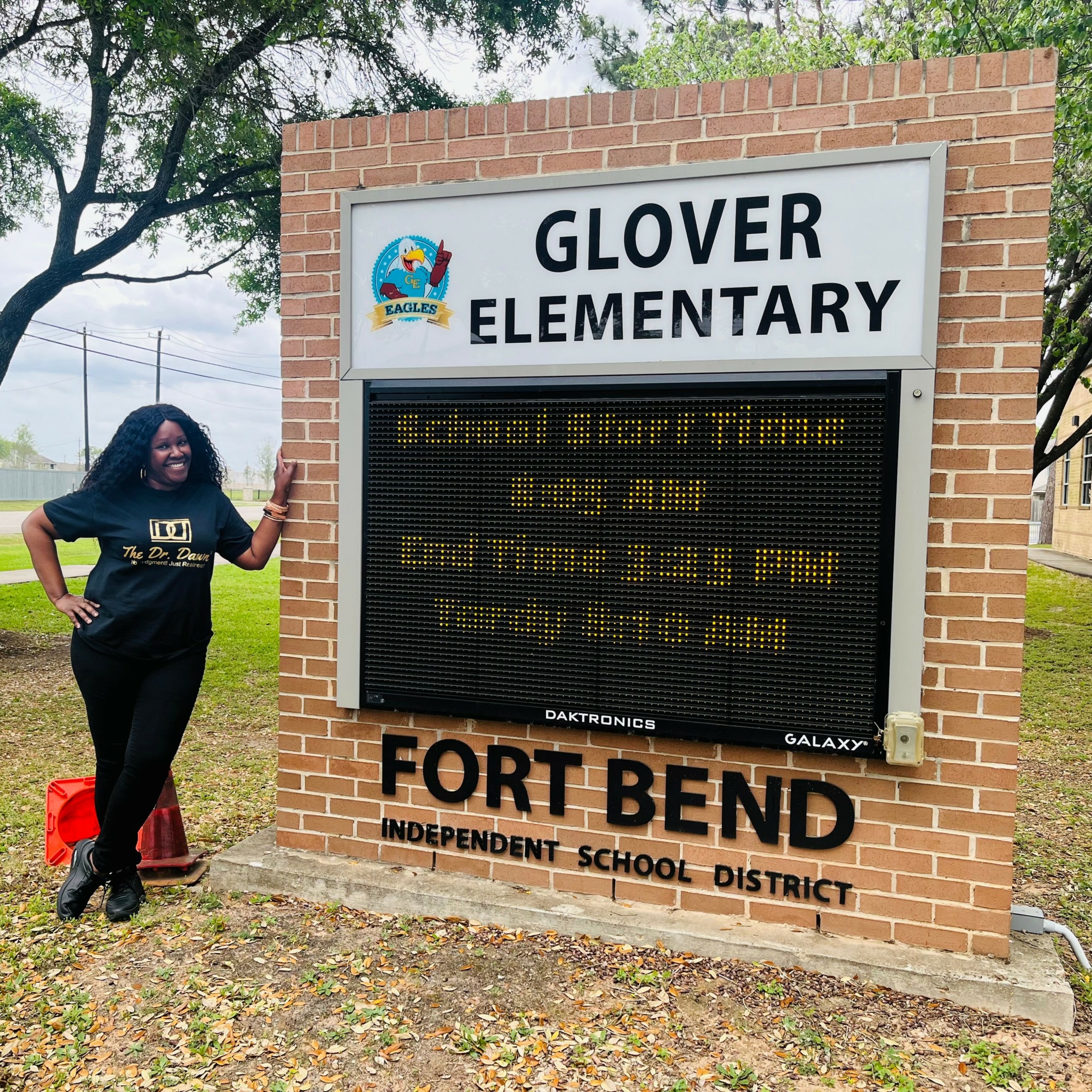Woman standing next to a brick sign for Glover Elementary School, smiling and pointing at the sign.