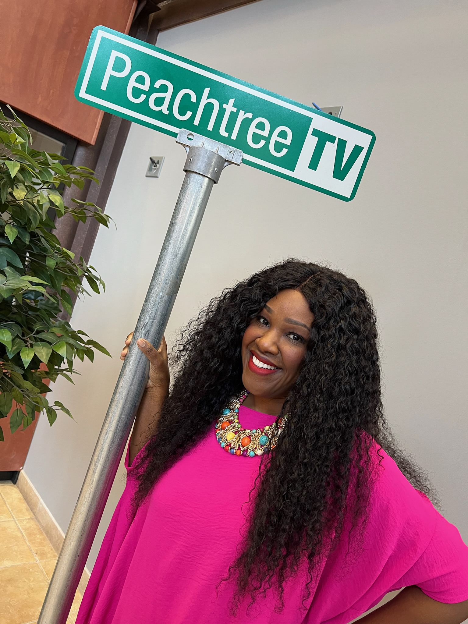 Woman with long curly hair wearing a pink top and colorful necklace, smiling near a street sign that reads Peachtree TV.