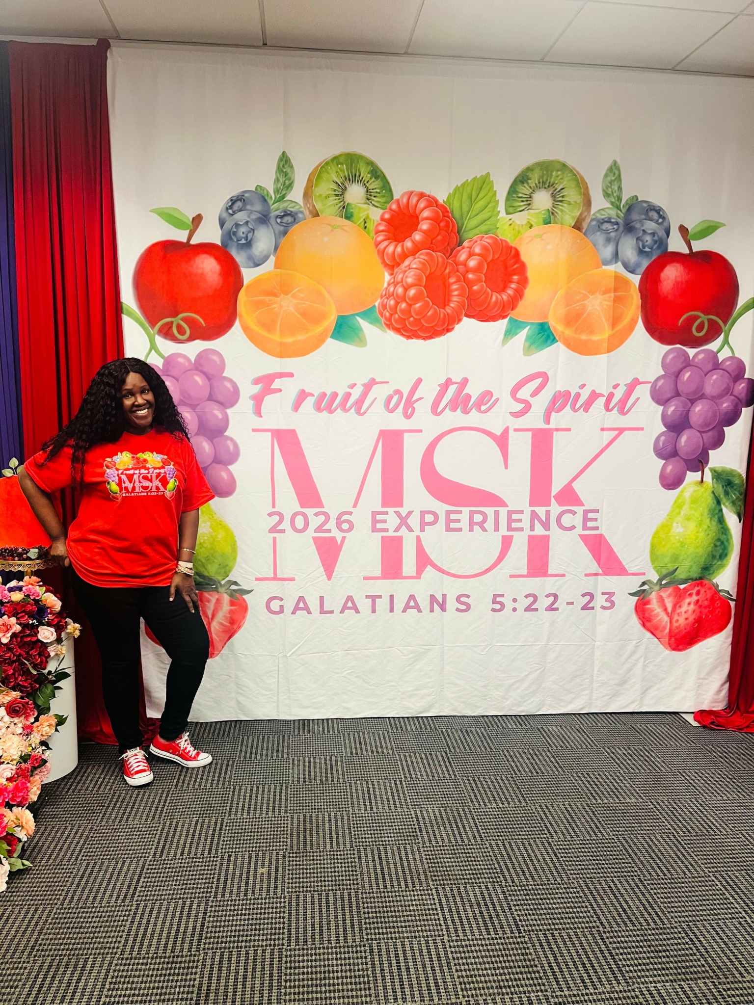 Woman standing in front of a colorful fruit-themed backdrop with text, smiling, wearing a red shirt and black pants.