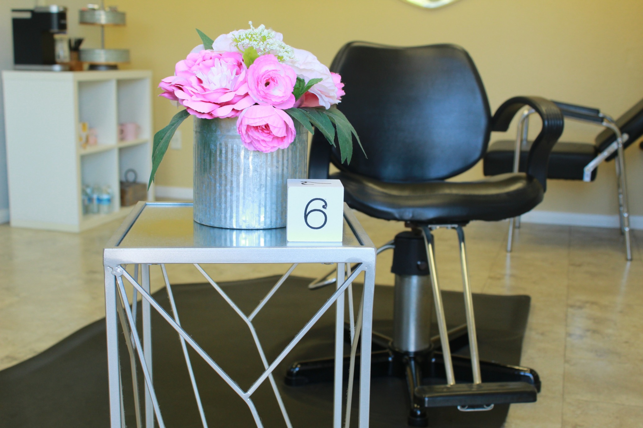 Flower arrangement in a metal container on a white side table, with a black chair in the background.