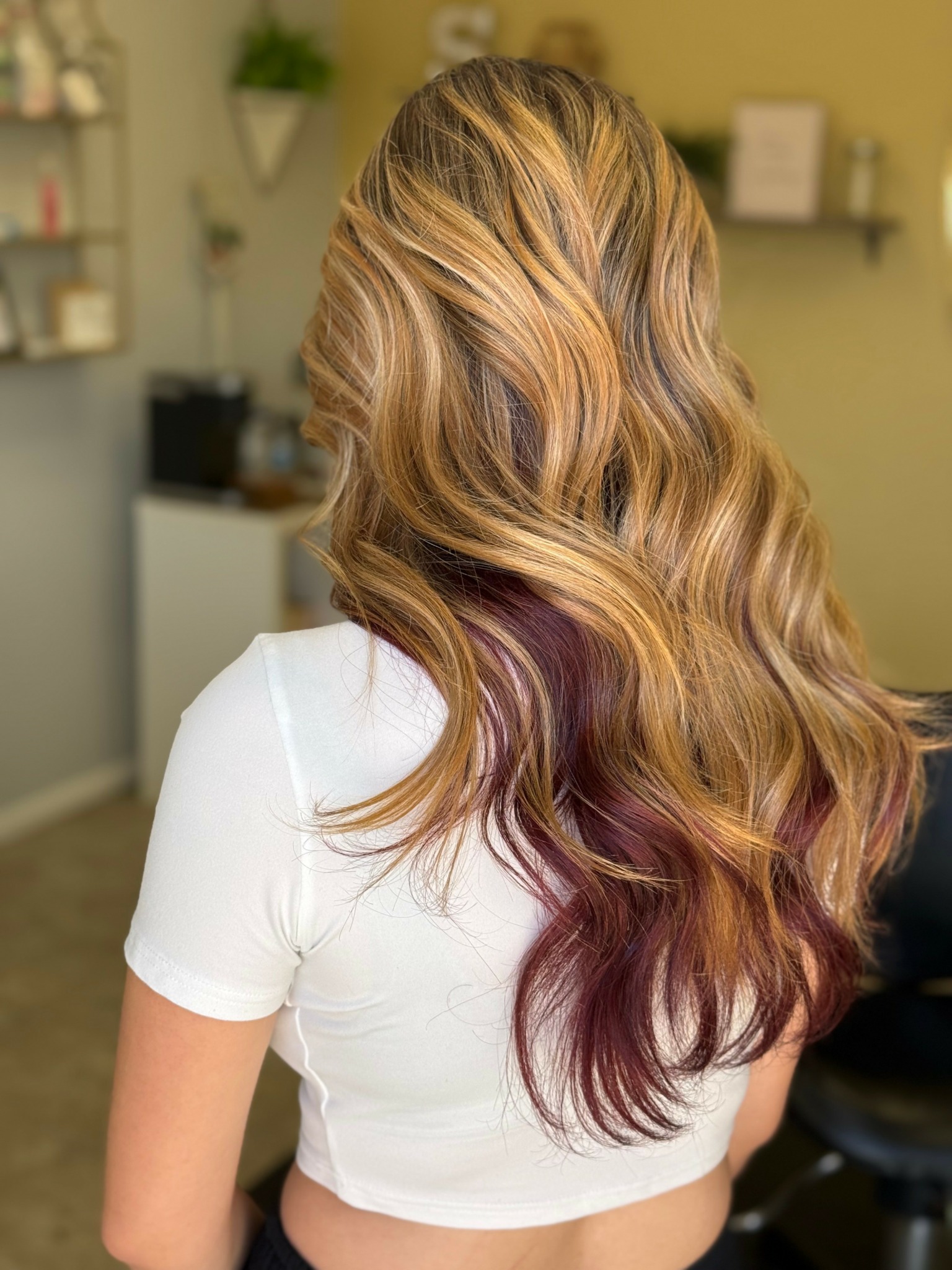 Back view of a woman with long, wavy hair with blonde and purple highlights, wearing a white top.