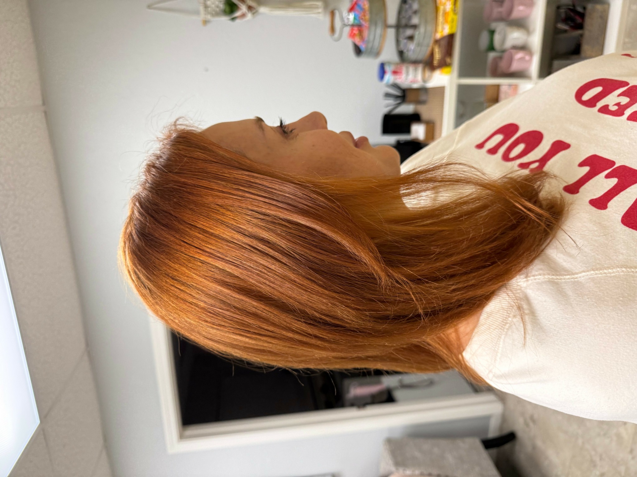 Side view of a person with long, reddish-brown hair in a room with shelves and various items.