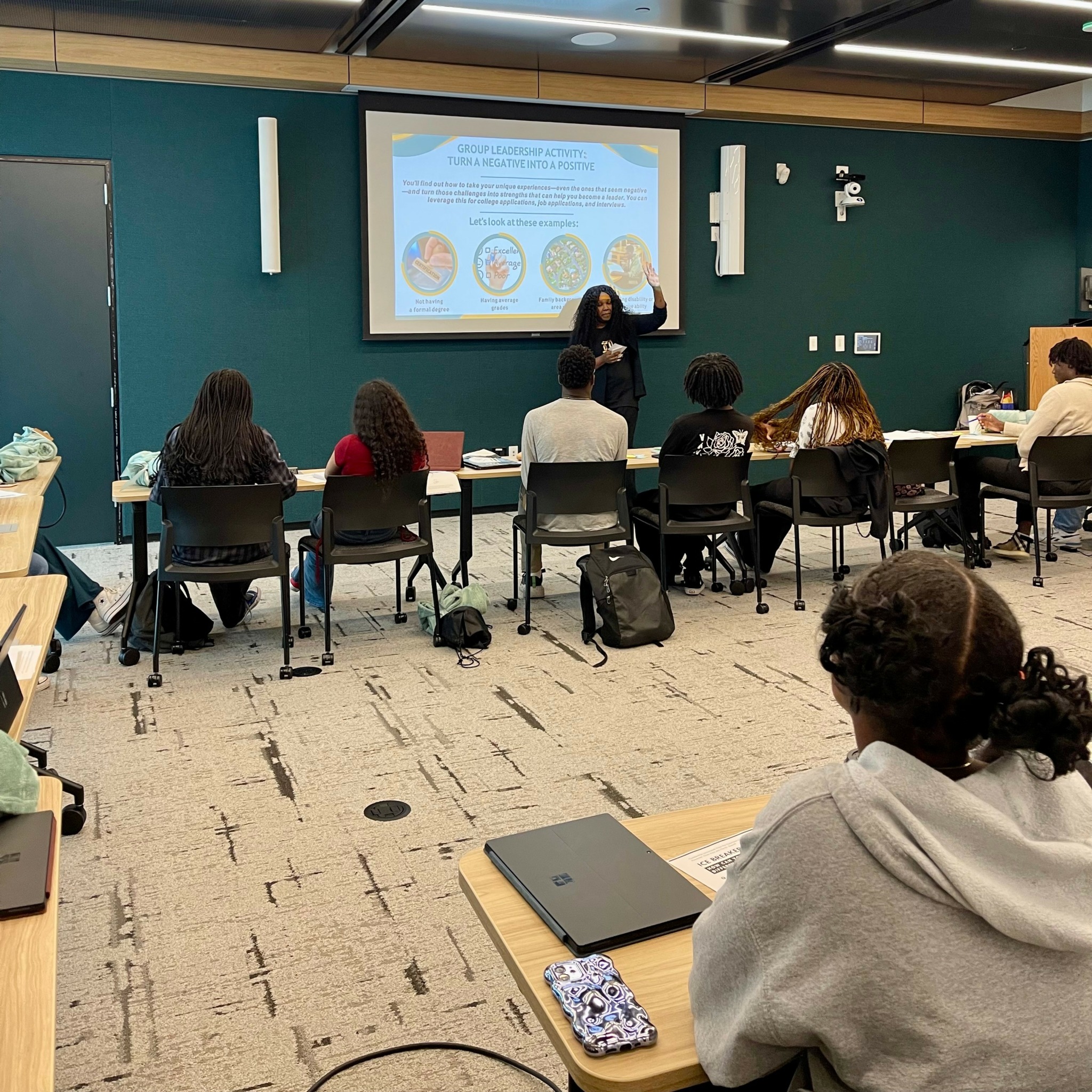 Classroom with students seated at desks facing a presenter and a projection screen, in a room with teal walls.
