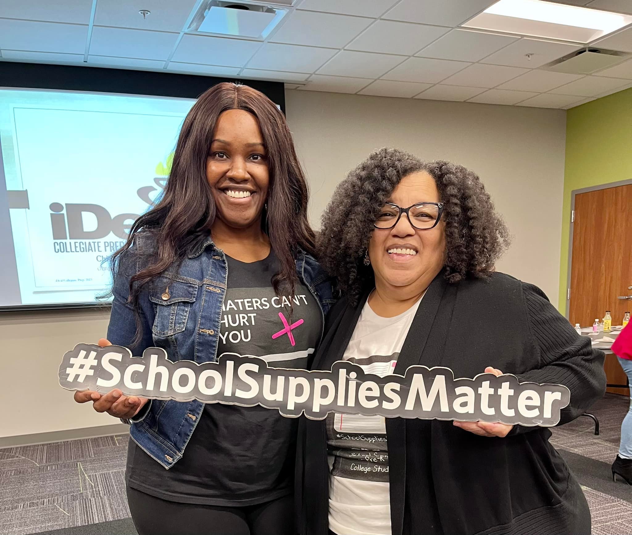 Two women smiling holding a sign that reads #SchoolSuppliesMatter in a room with a presentation screen.