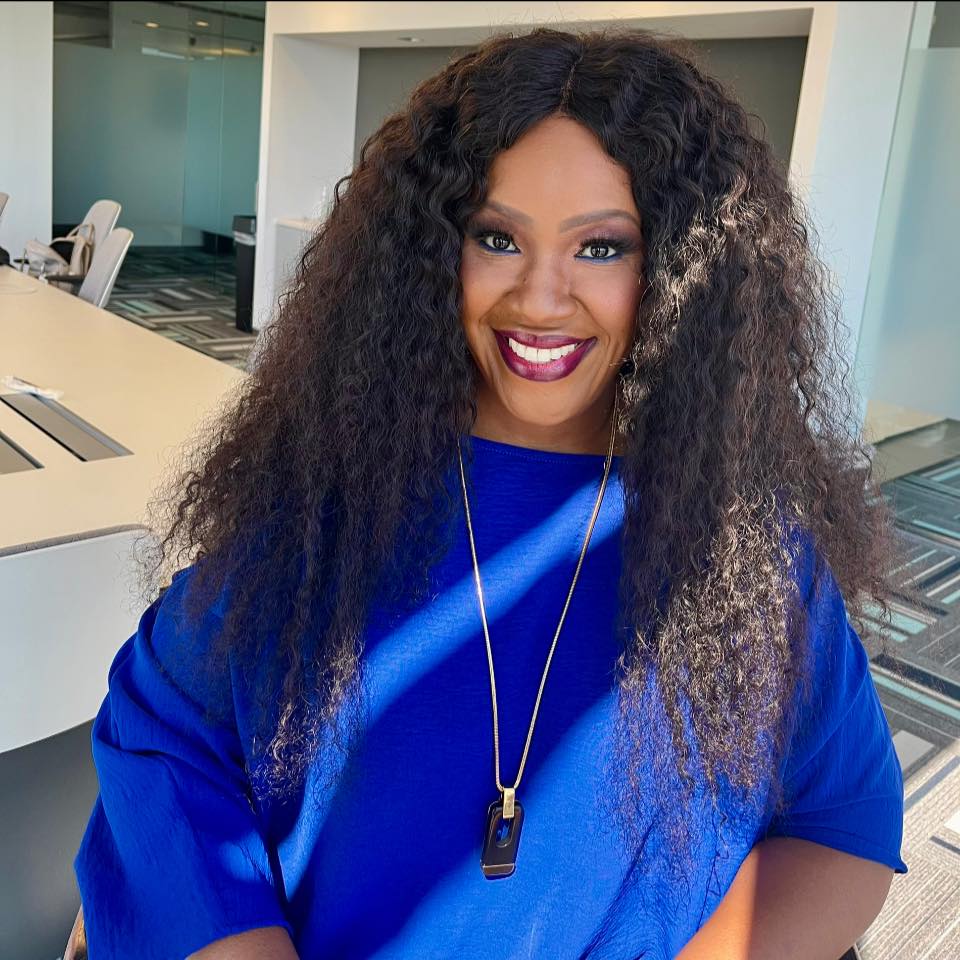 Woman with curly hair smiling, wearing a blue top and necklace, in a modern office space.