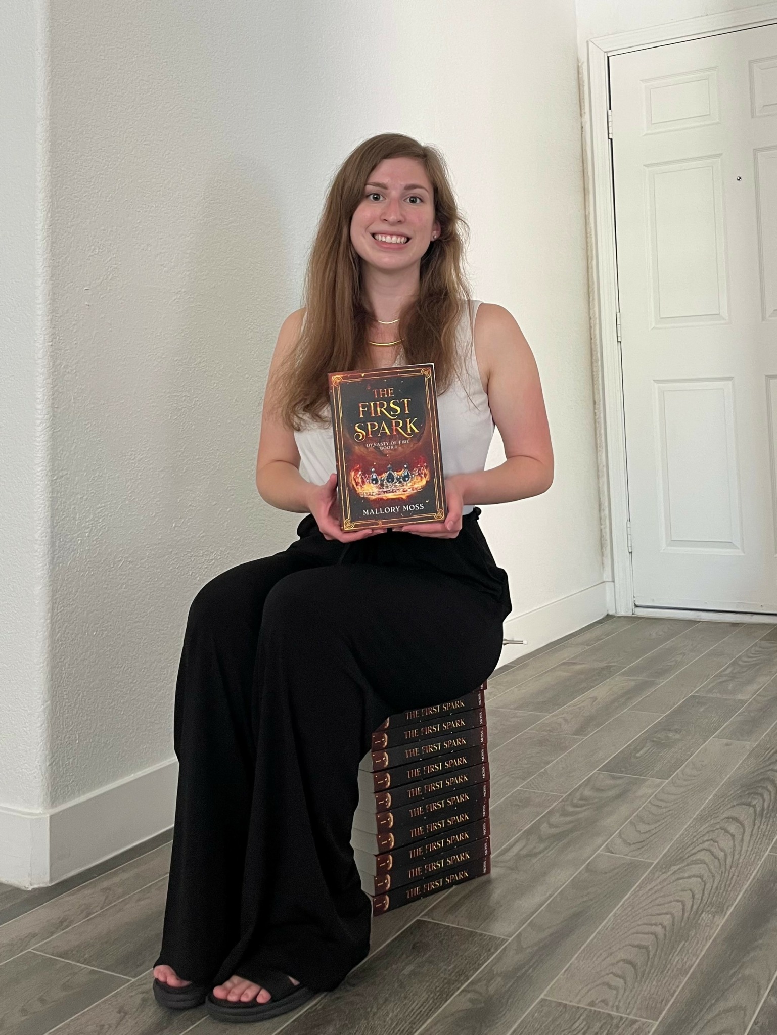 Young woman with long hair sitting on a box, holding a book, smiling, in a room with white walls and wooden floor.