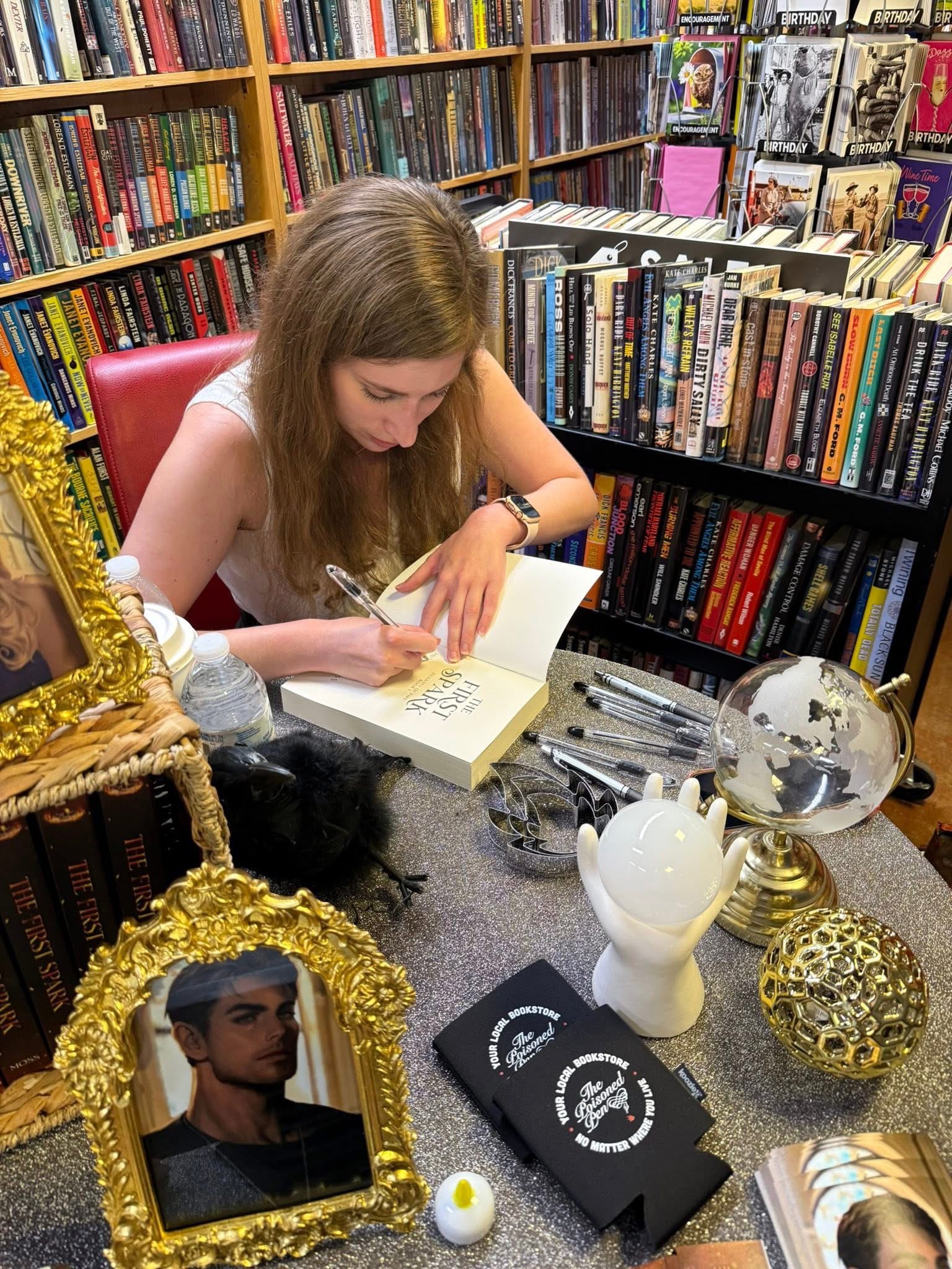 Young woman with long hair reading and writing in a book at a table in a bookstore, surrounded by books and decorative items.