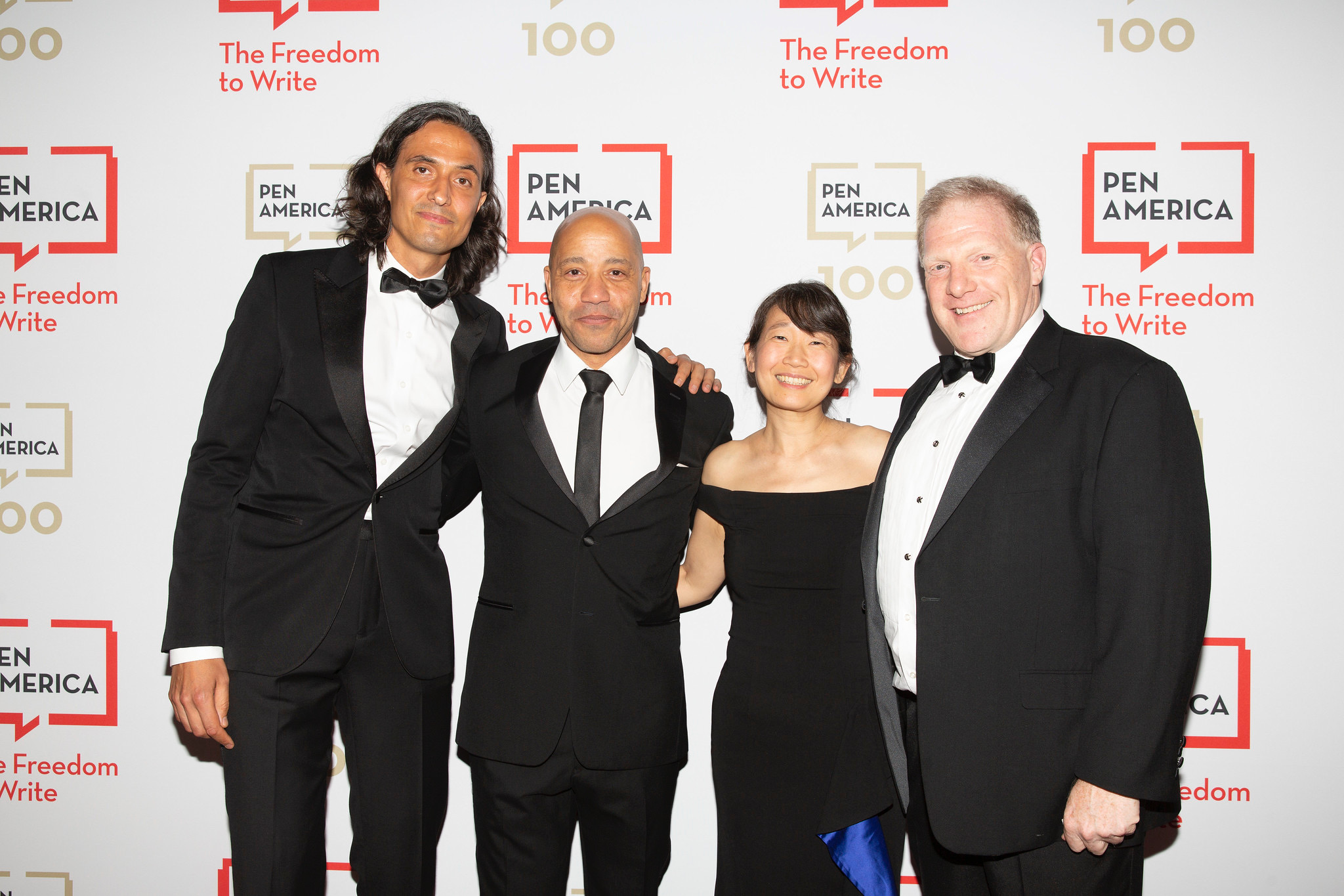 Four people in formal attire standing together at an event with a backdrop featuring logos and text.