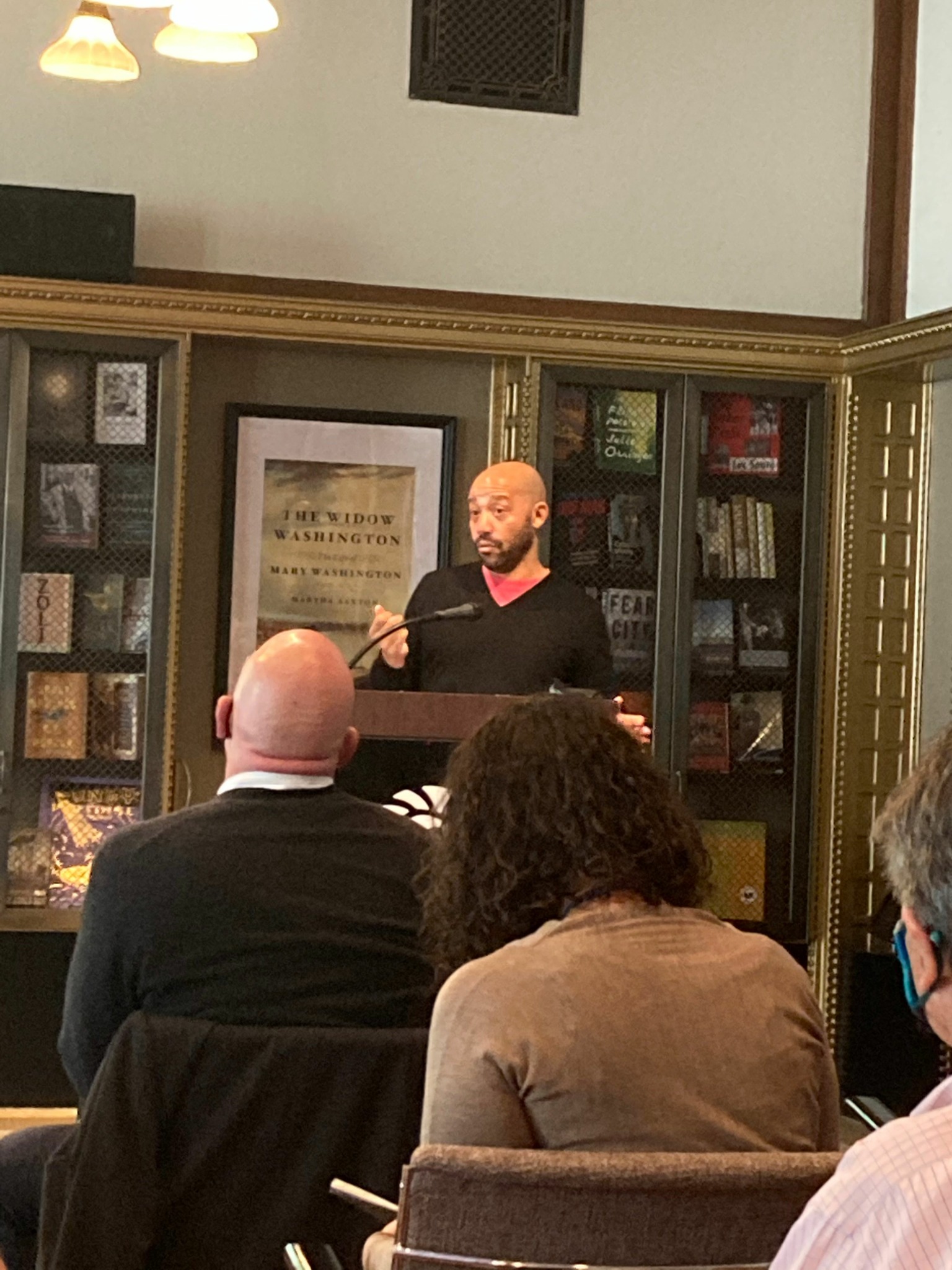 Person speaking at a podium in a room with bookshelves and framed pictures, audience members seated facing the speaker.