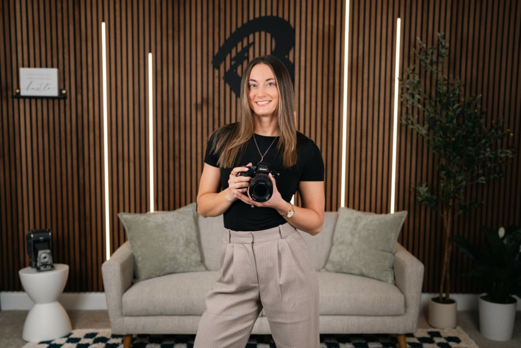 Woman holding camera, smiling, sitting on a light-colored sofa with pillows, in a room with wooden wall and plants.