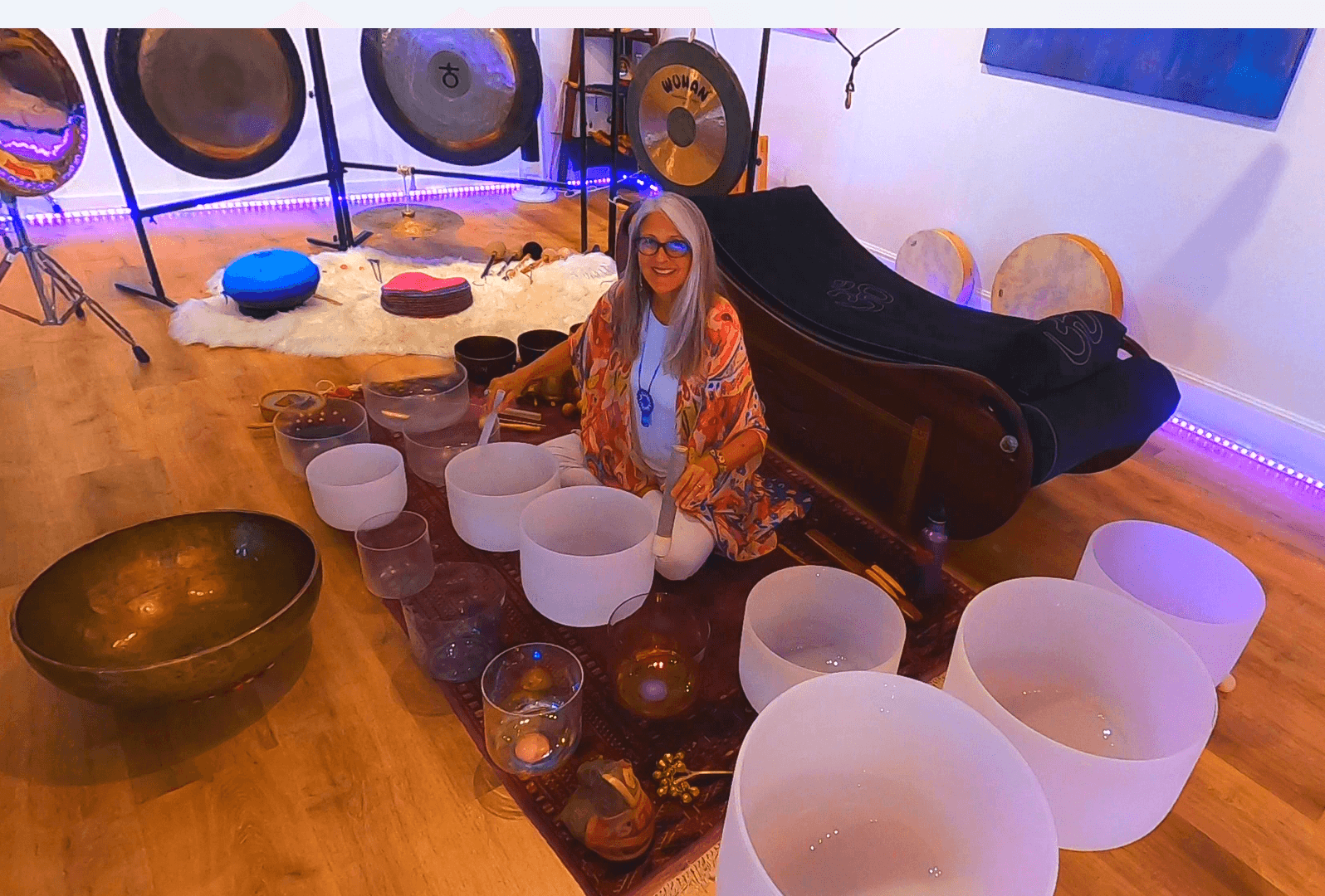 Person with long hair and glasses sitting on the floor surrounded by singing bowls and white chairs.