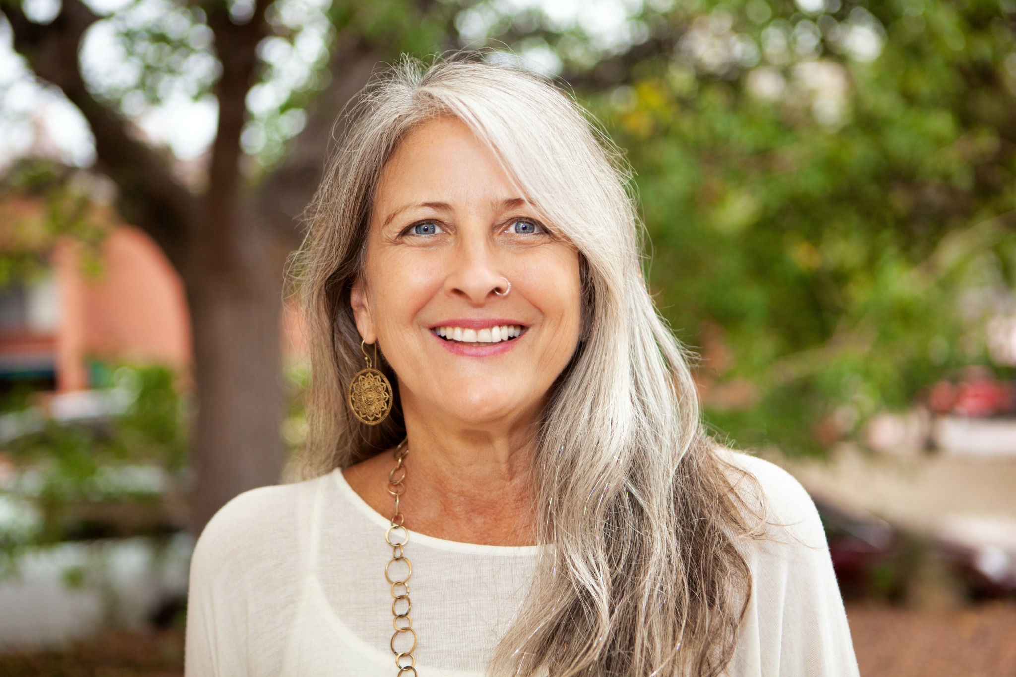 Smiling woman with long gray hair outdoors, trees and blurred background behind her.