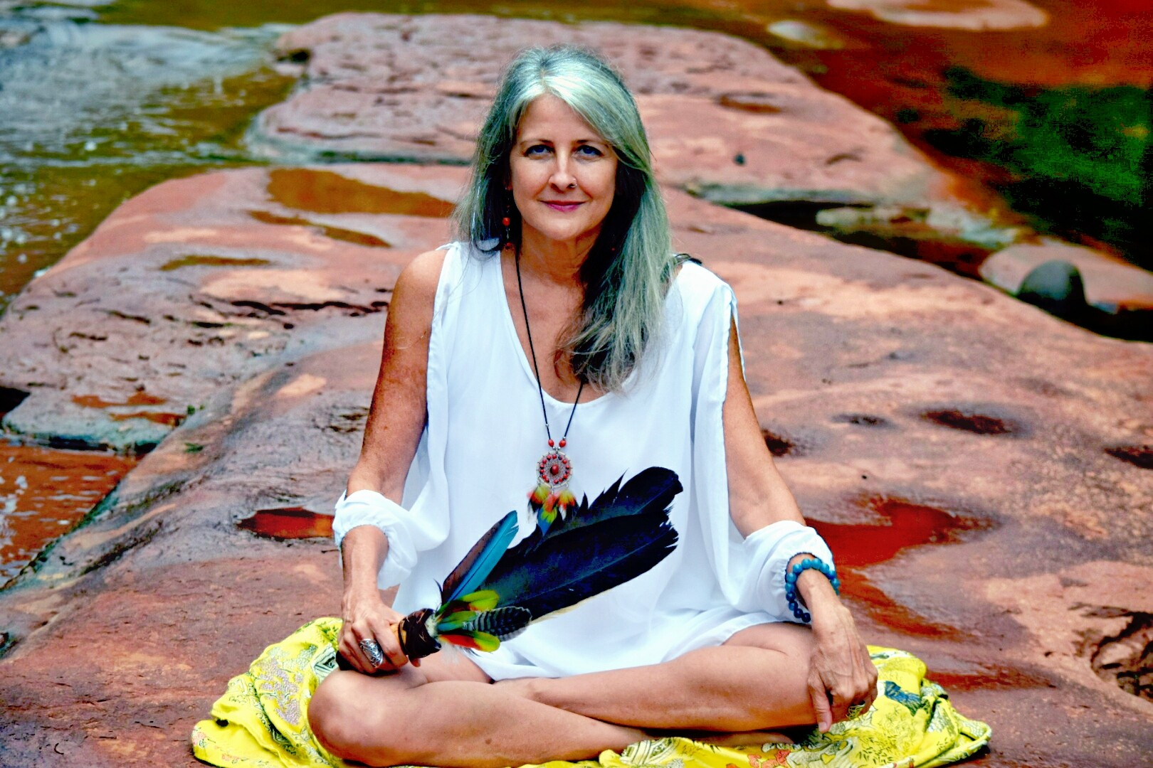 Woman sitting cross-legged on rocks near water, holding feathers, smiling, wearing white dress and jewelry.