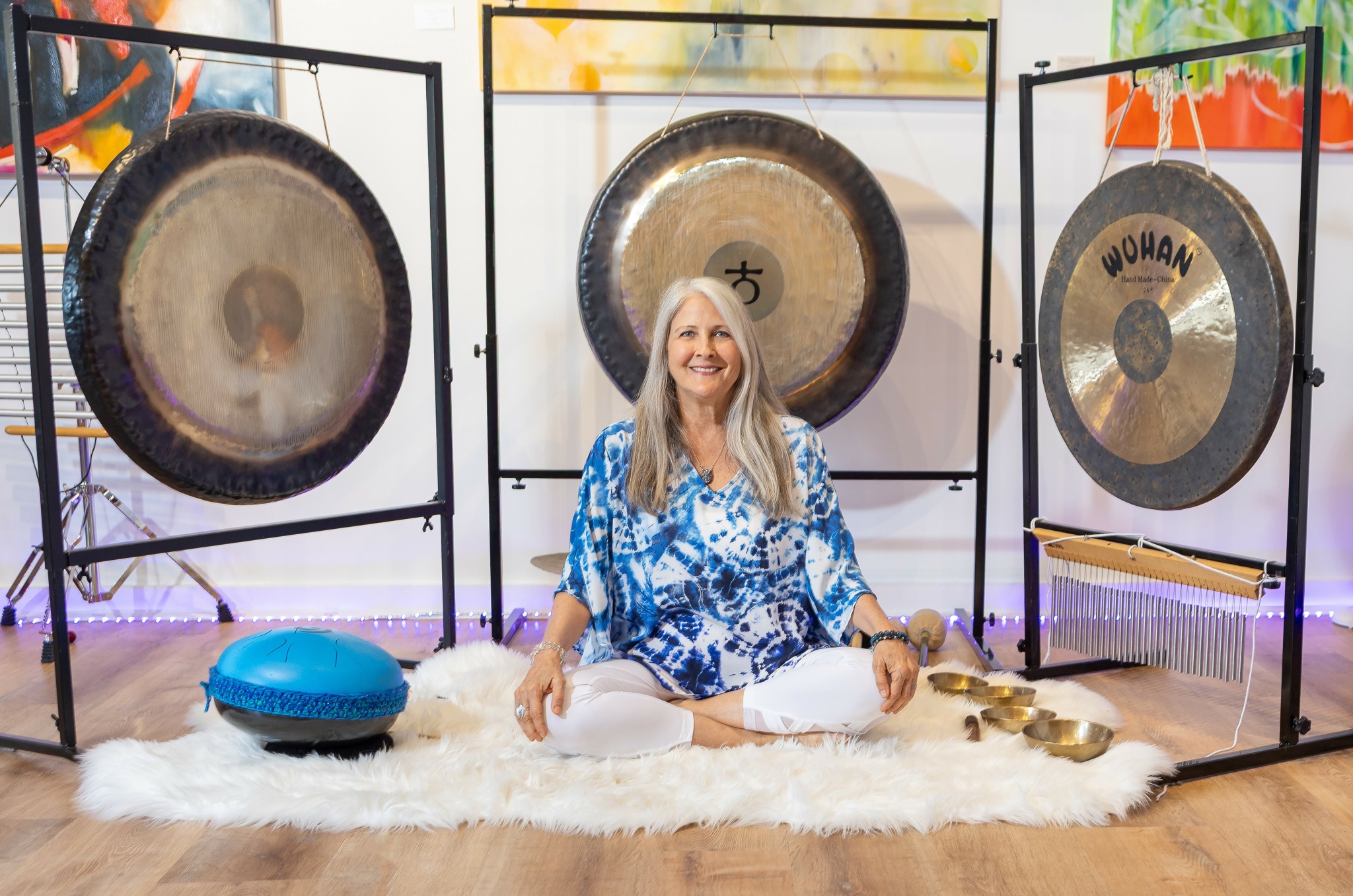 Woman sitting on a rug with three large gongs behind her and a blue cushion nearby.