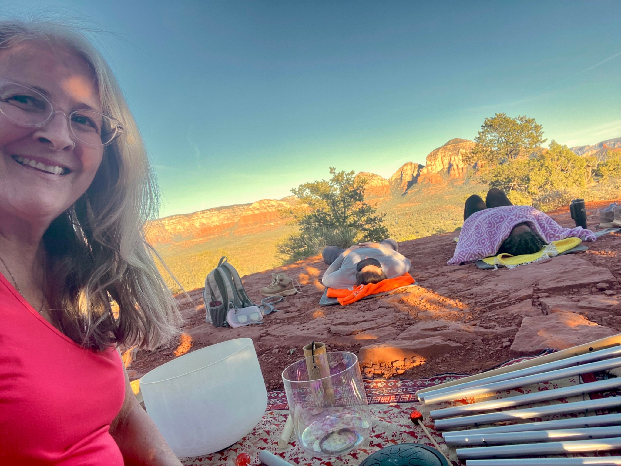 Woman smiling outdoors with a desert landscape, rocks, and people lying on the ground in the background.