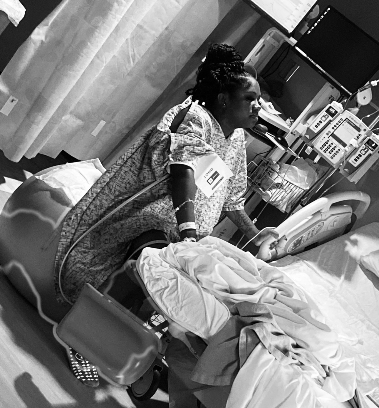 Woman in hospital gown sitting on bed with medical equipment around her, holding a phone, in a hospital room.