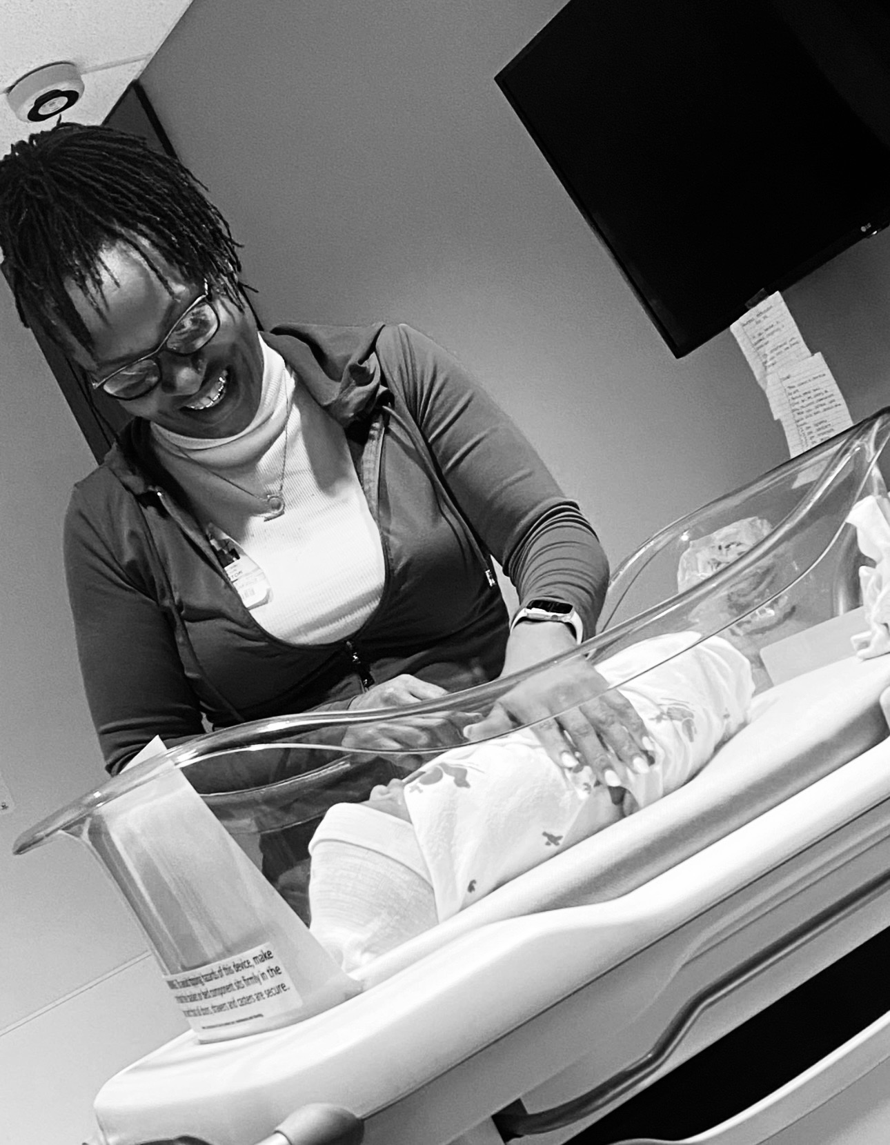 Woman smiling at a newborn in a hospital bassinet, wearing glasses and a jacket.