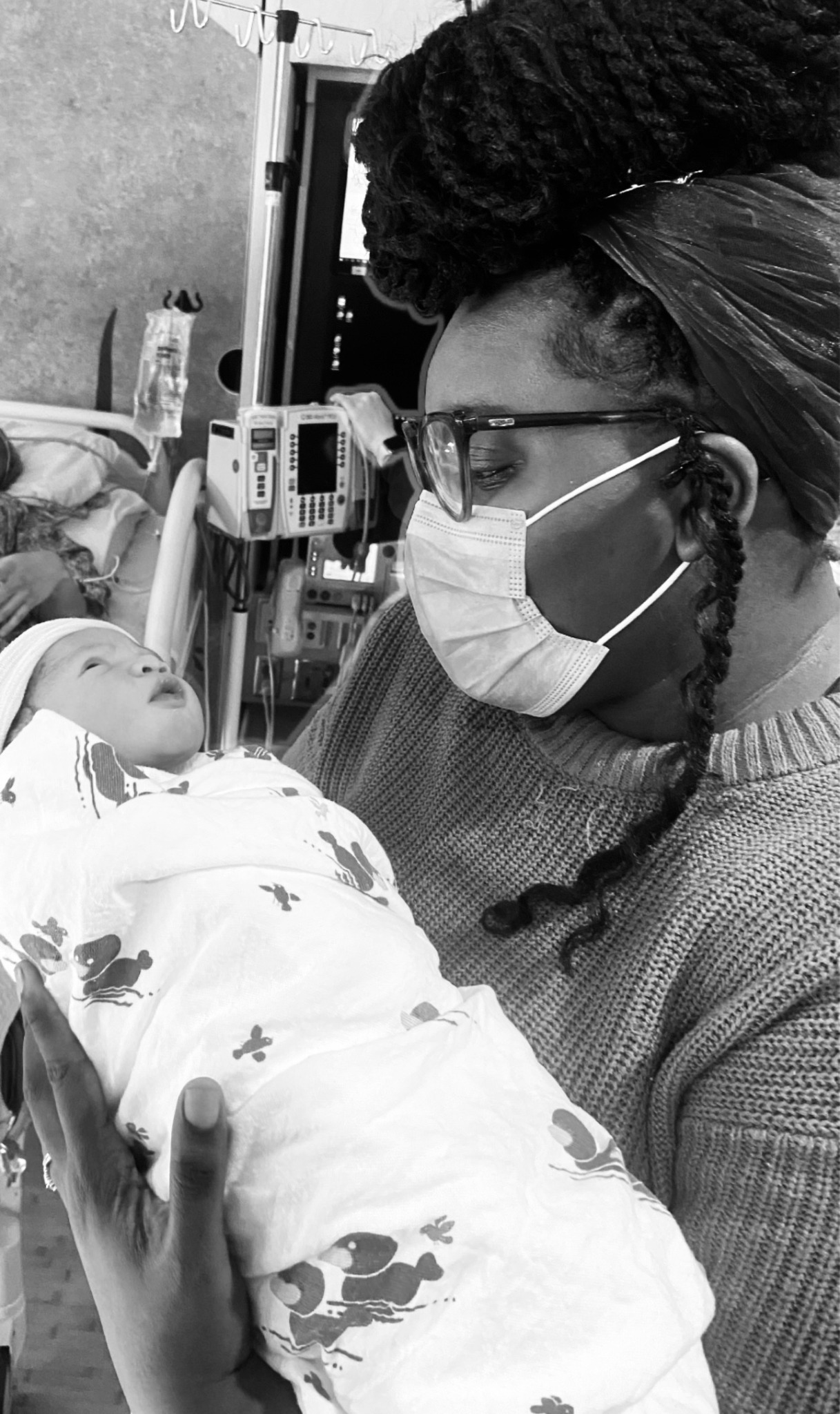 Woman wearing glasses and a face mask holding a baby wrapped in a blanket.