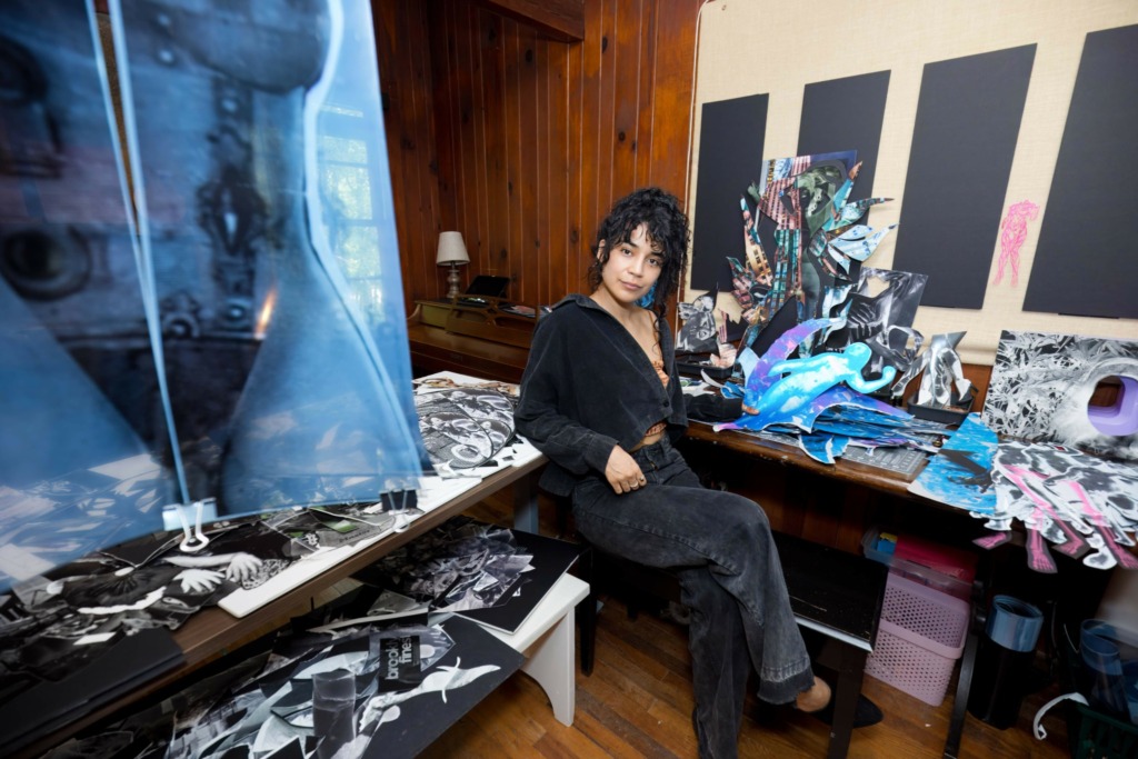 Woman with curly hair in black outfit sitting at cluttered desk with fabric and art supplies in a room with wooden walls.