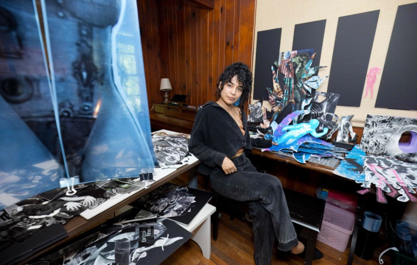 Woman with curly hair in black outfit sitting at cluttered desk with fabric and art supplies in a room with wooden walls.