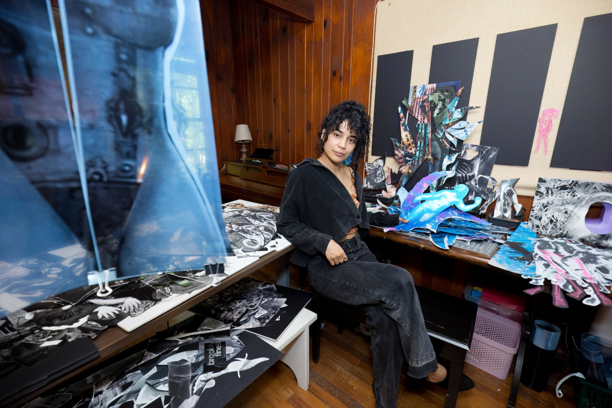 Woman with curly hair in black outfit sitting at cluttered desk with fabric and art supplies in a room with wooden walls.