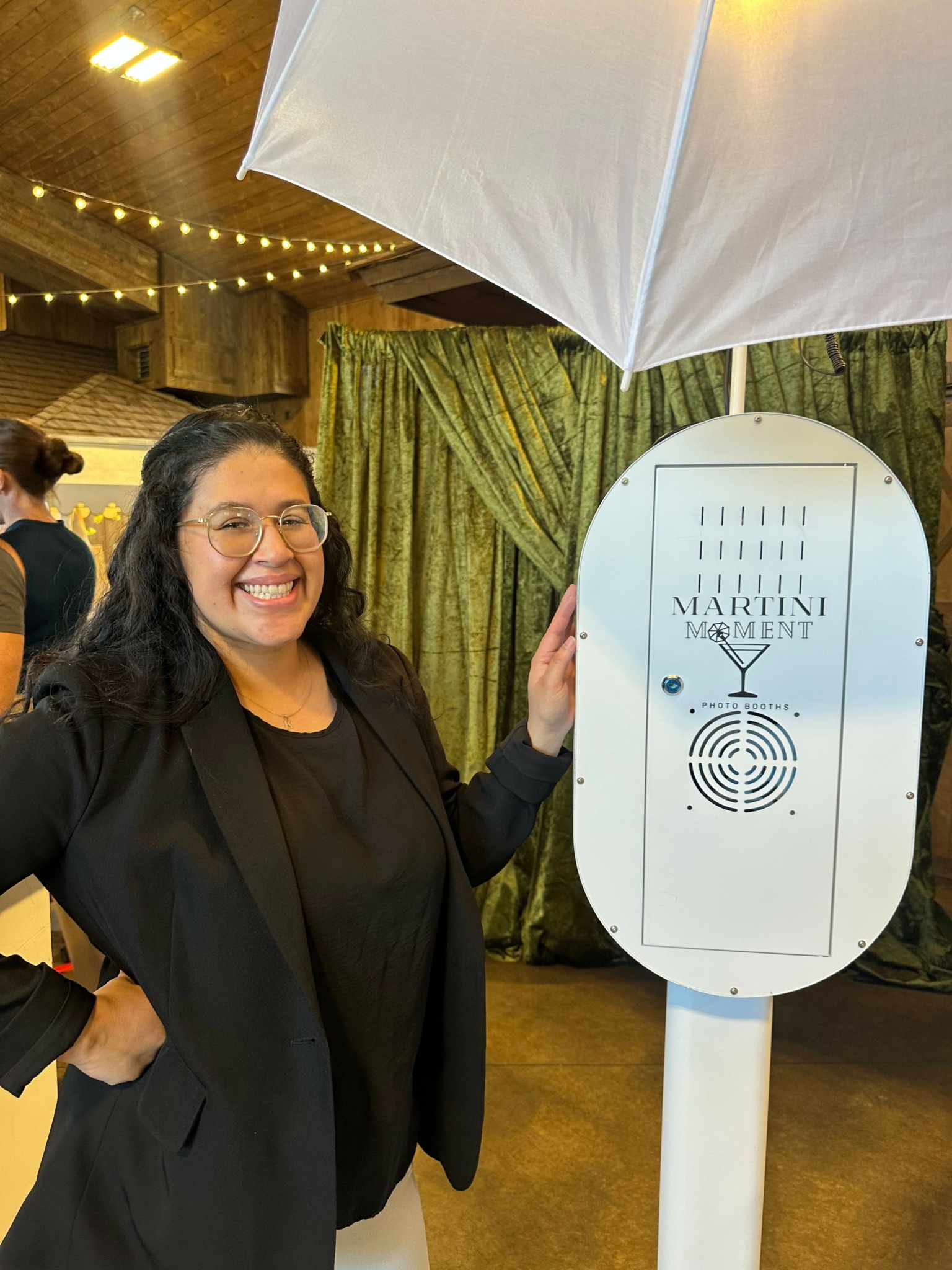 Woman smiling, standing indoors near a white umbrella and a tall white clock with black markings.