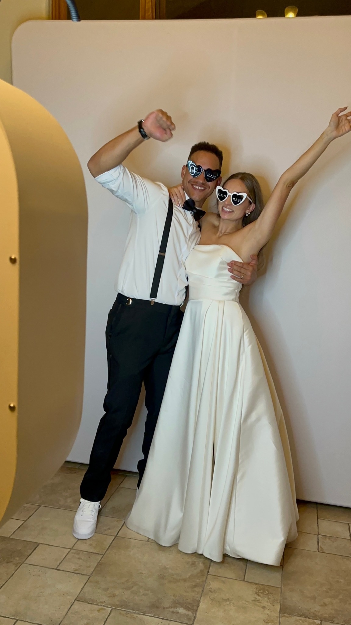 A man and woman in formal attire pose together indoors, smiling and wearing sunglasses, with the woman raising her arm.