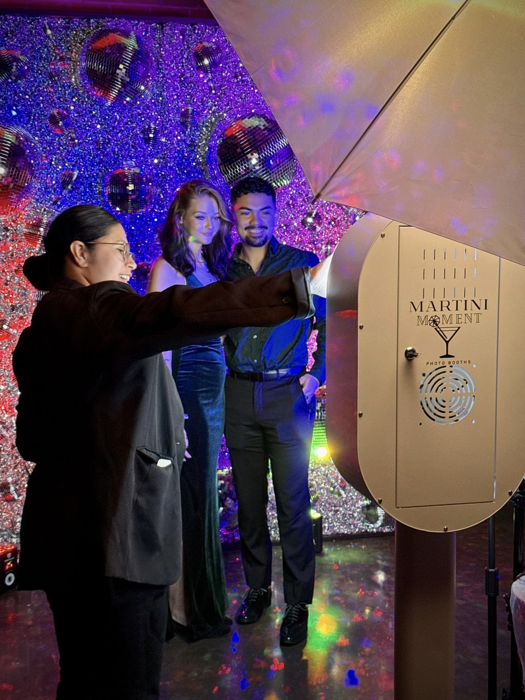 Three people standing near a large champagne dispenser at a celebration event, with colorful decorations in the background.