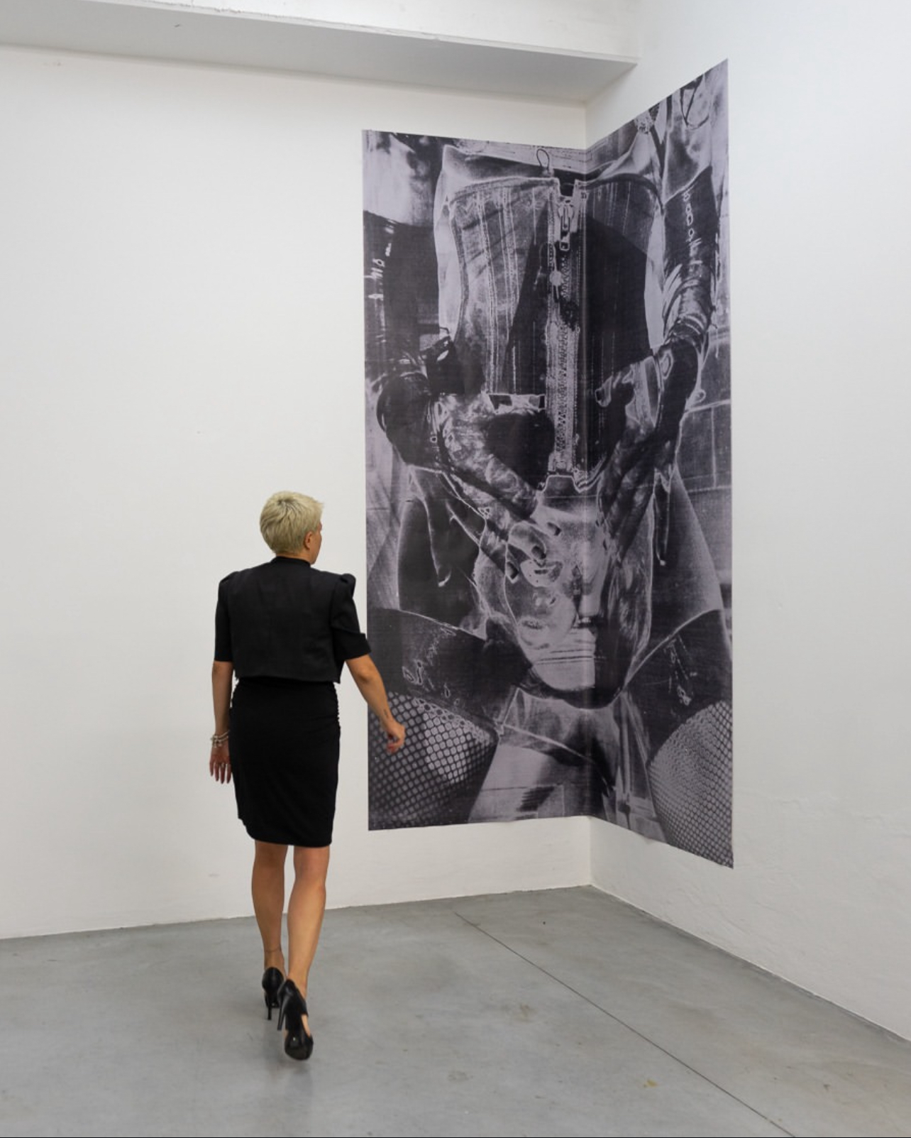 Woman with short blonde hair in black dress walking in art gallery, looking at large black-and-white abstract artwork on wall.