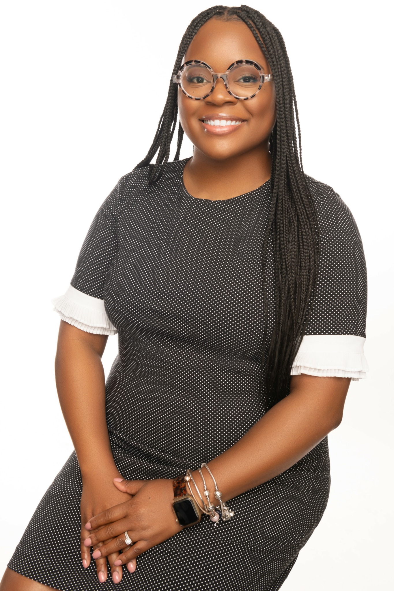 Smiling woman with glasses and long braided hair, wearing a black dress with white cuffs, standing with hands clasped.