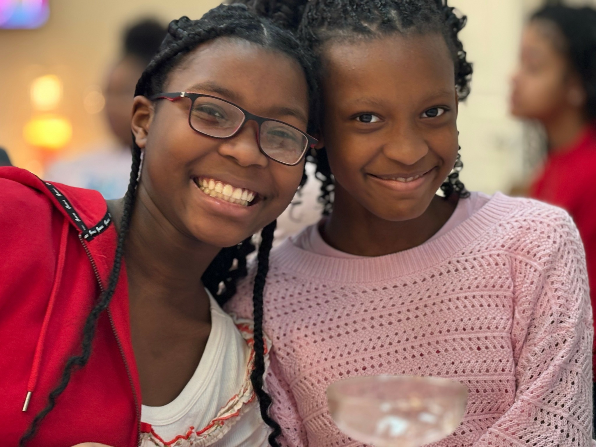 Two smiling girls with braided hair, one wearing glasses and a red jacket, the other in a pink sweater, in a social setting.