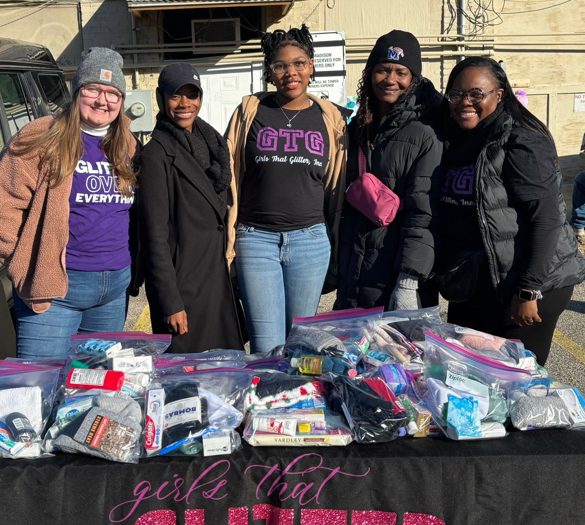 Five women standing behind a table with bags of supplies outdoors, smiling, wearing casual clothing and beanies.